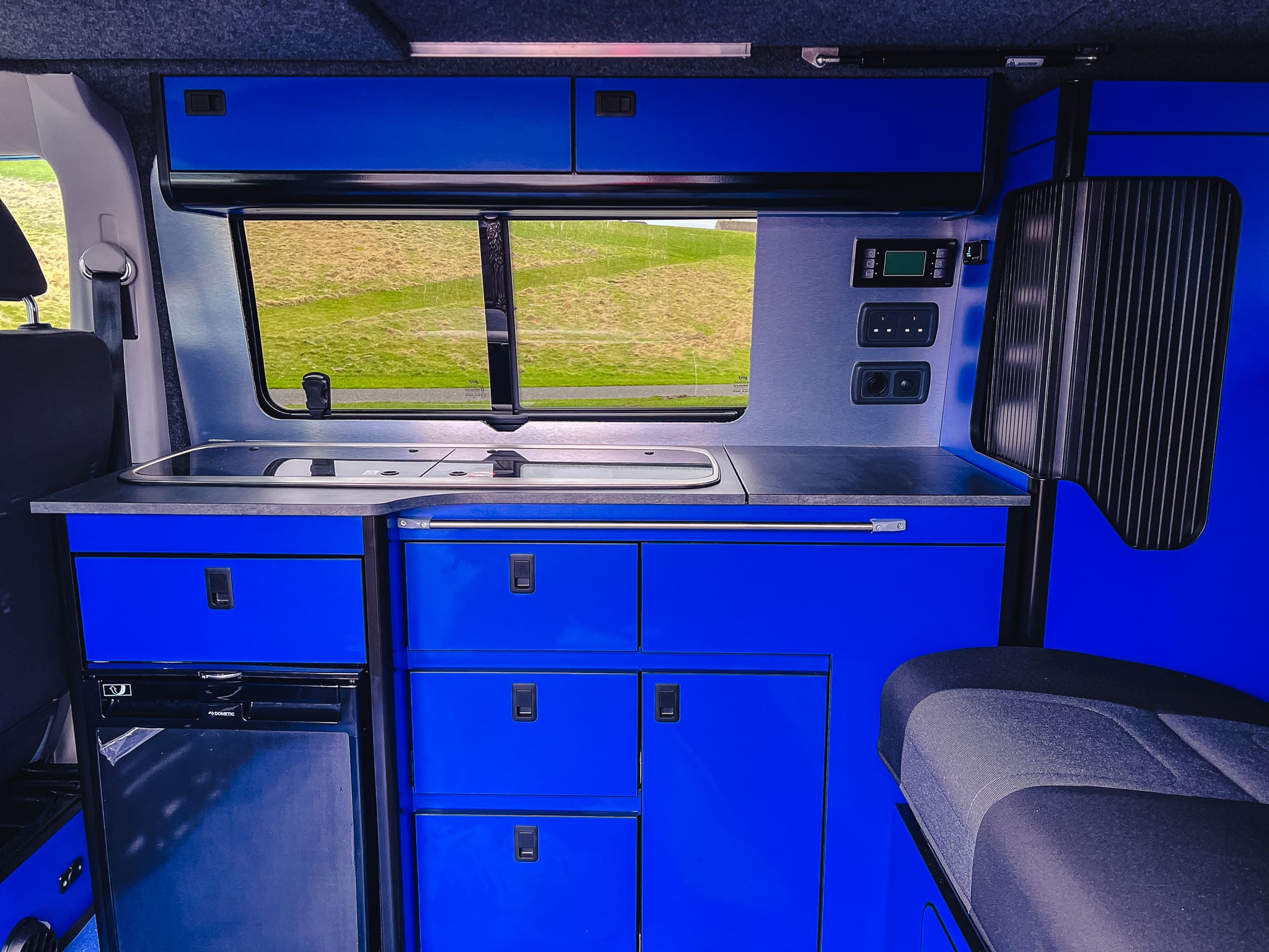 Interior of a rental camper van with bright blue cabinets, a compact sink, drawers, mini fridge, control panels, and a window revealing Scotland’s grassy landscape. The motorhome setup is modern and space-efficient.