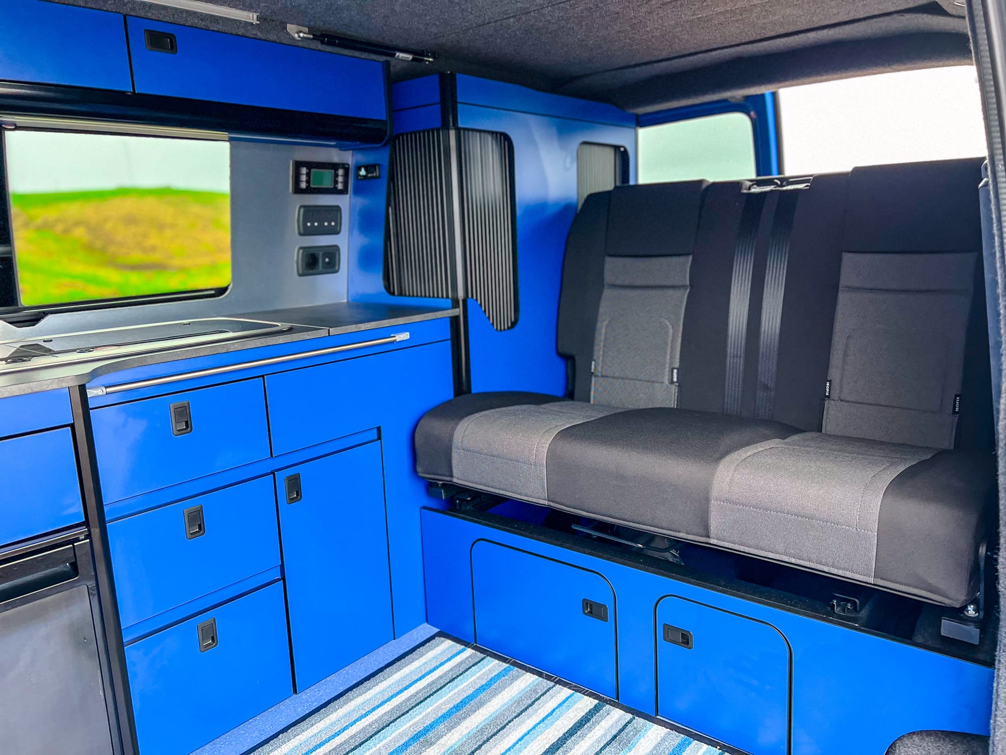 Interior of a camper van rental near Peterhead, Scotland, featuring bright blue cabinets, a small sink, control panels, a striped rug, and a black and gray bench seat with storage. A window reveals green grass outside.