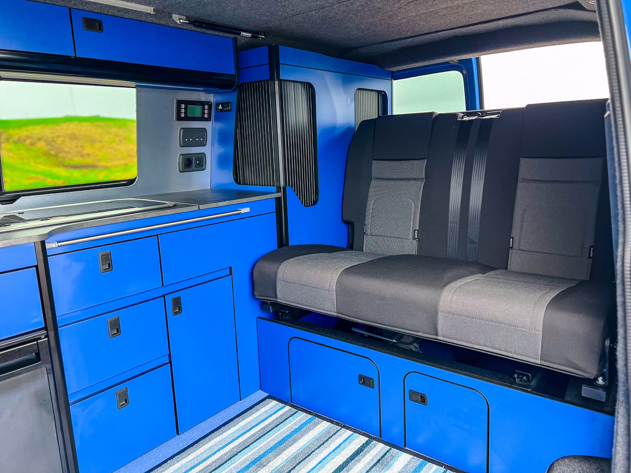 Interior of a camper van rental near Peterhead, with bright blue cabinets, a small sink, control panels, and a gray two-seat bench. The floor has a striped rug, and the window reveals green fields reminiscent of Scotland’s charm.