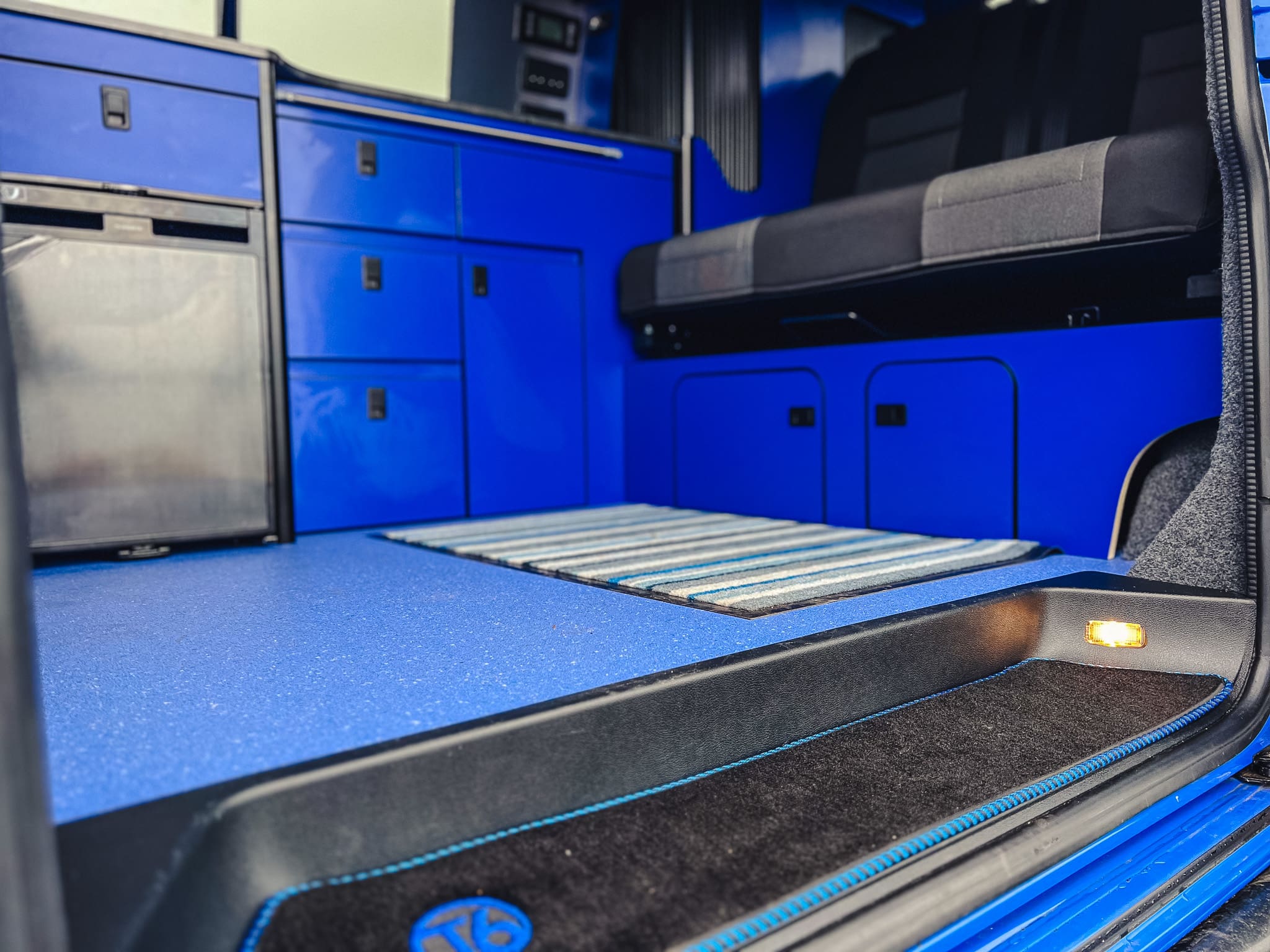 Interior of a blue camper van rental near Peterhead, Scotland, featuring cabinets, a small fridge, a striped rug, a bench seat, and ambient floor lighting—all designed with modern finishes and built-in storage.