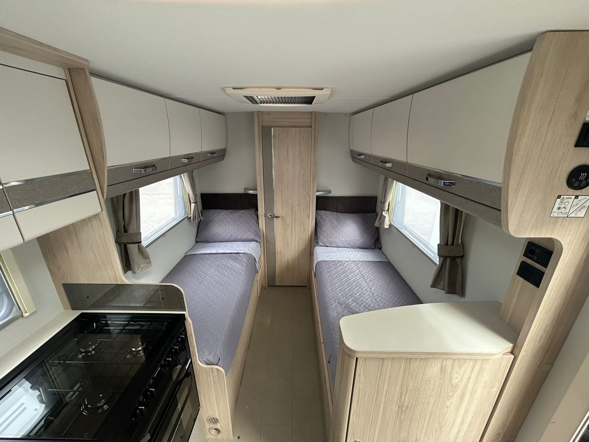 The interior of a modern motorhome rental with two single beds on either side, grey bedding, wood cabinets, windows with curtains, and a compact kitchen area with a stove in the foreground—perfect for exploring Peterhead.
