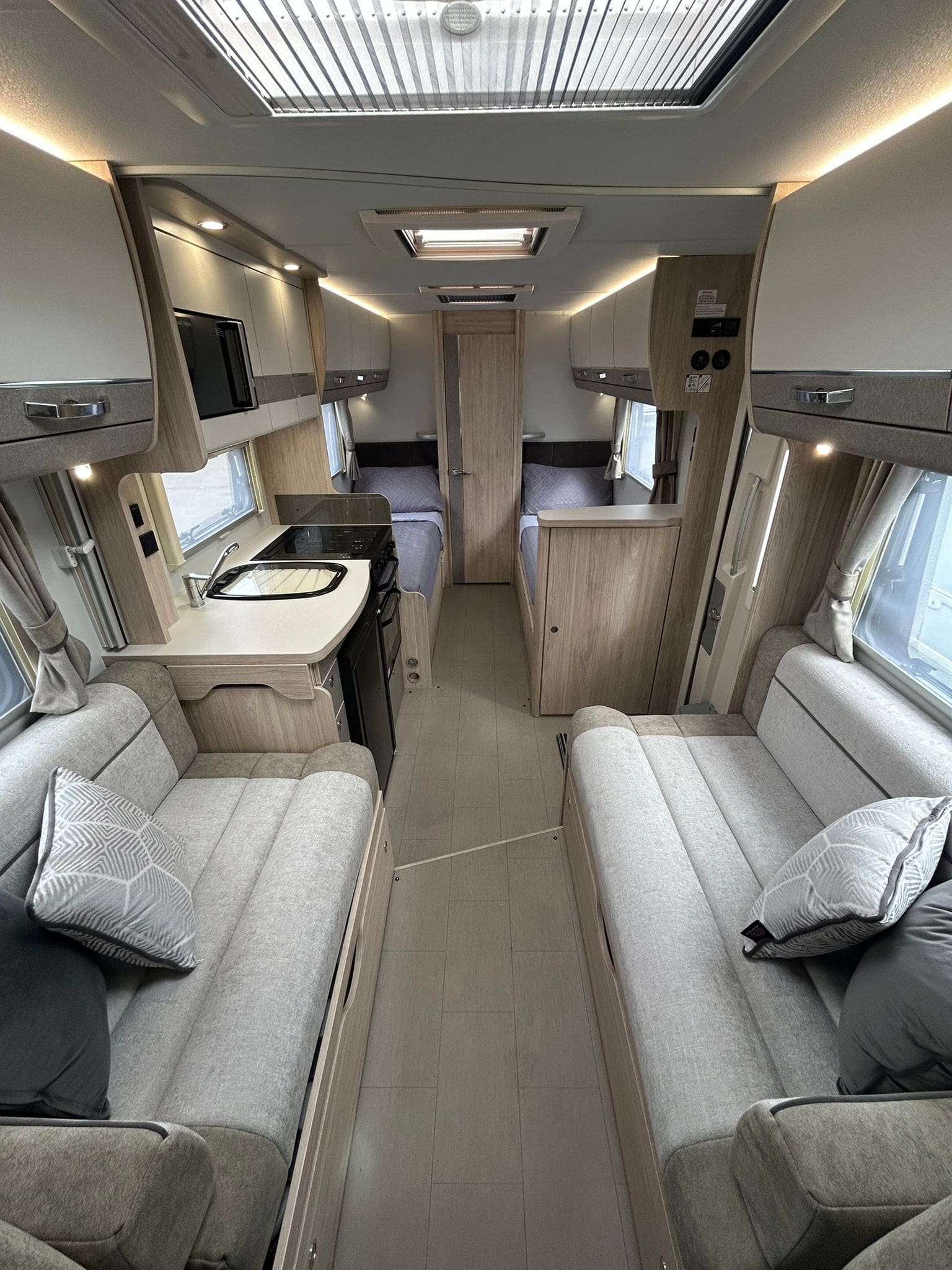 The interior of this modern motorhome rental features two gray sofas, a compact kitchen with stove and sink, overhead cabinets, and twin beds at the back—all in a bright, neutral color scheme perfect for exploring Scotland.