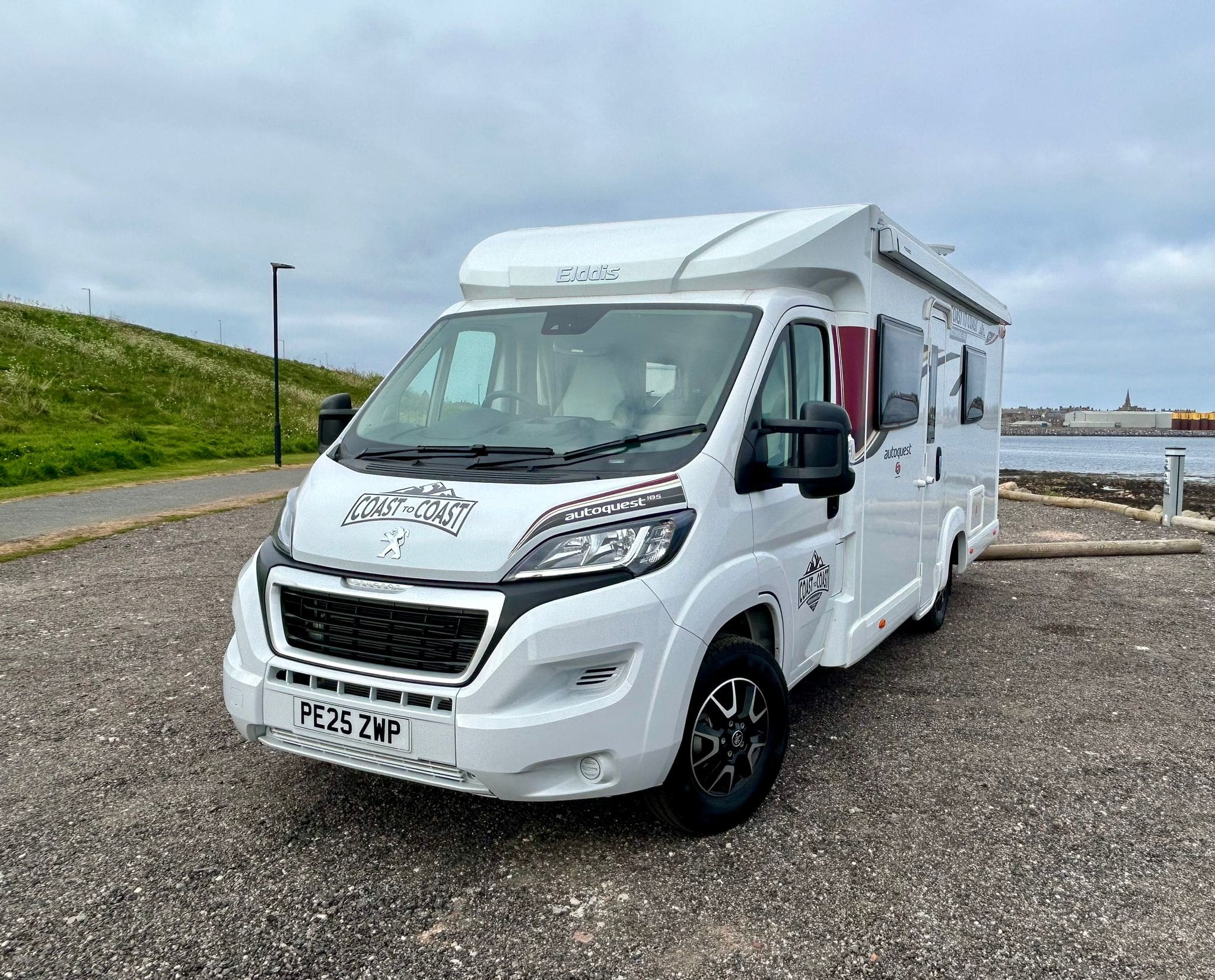 A white motorhome rental with a Coast to Coast decal is parked on a gravel area near the coast in Scotland, with green grass, a road, and water in the background under a cloudy sky.