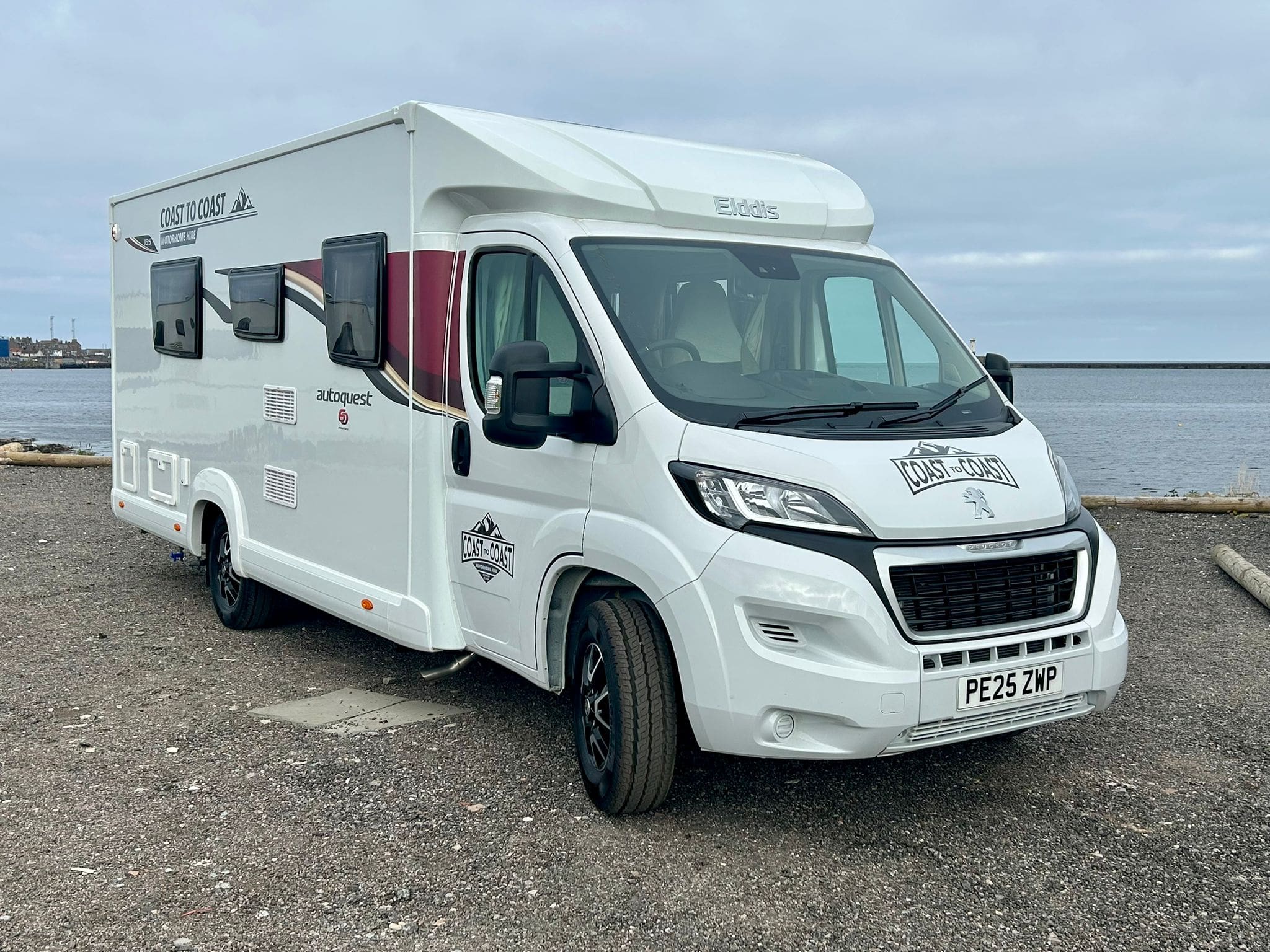 A white Peugeot motorhome with maroon and grey detailing is parked on gravel near the Peterhead waterfront on an overcast day. The vehicle has a UK license plate and “Autoquest” branding on the side and front.