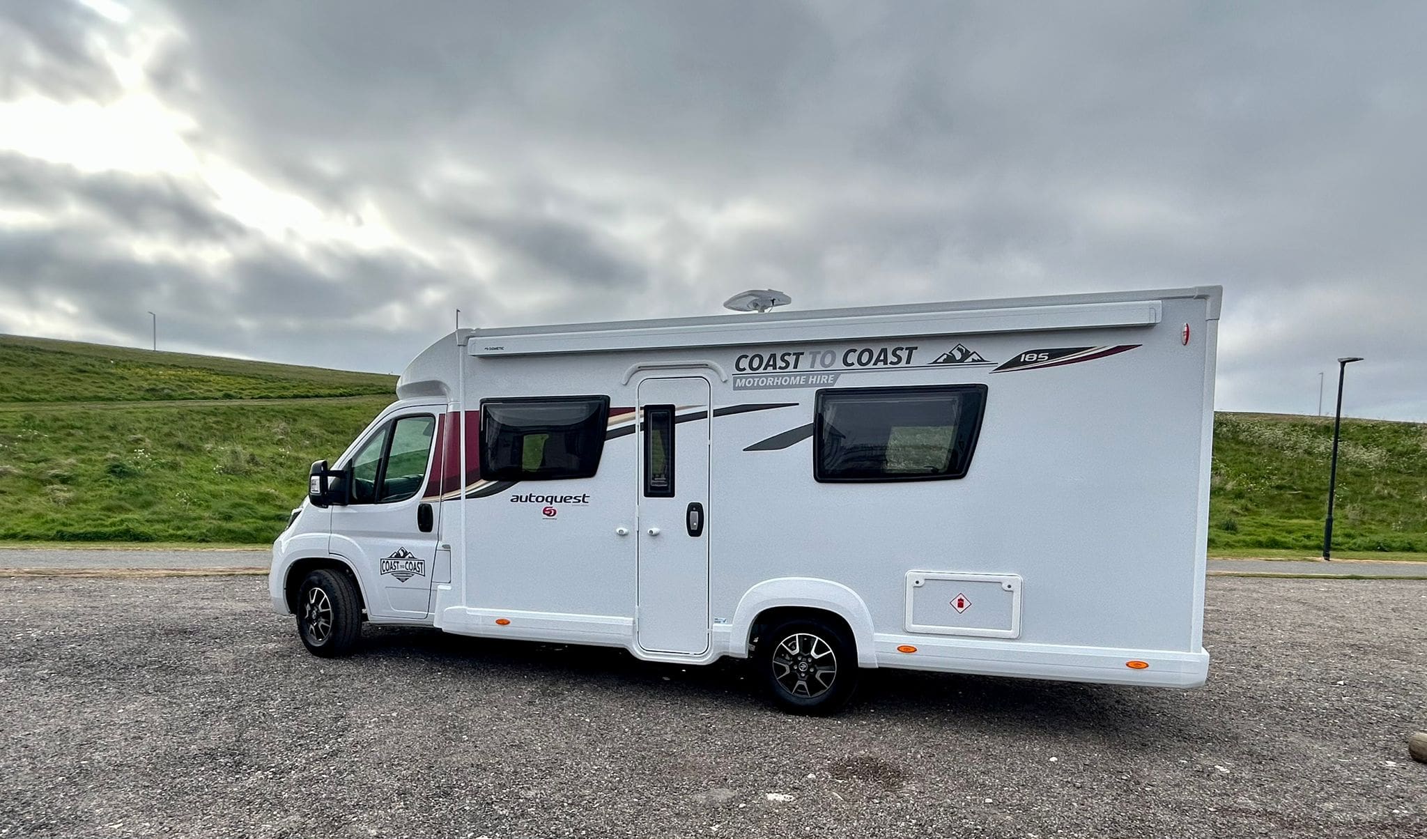 A white Coast to Coast rental motorhome is parked on a gravel lot near Peterhead, with green grassy hills and a cloudy sky in the background, capturing the scenic beauty of Scotland.