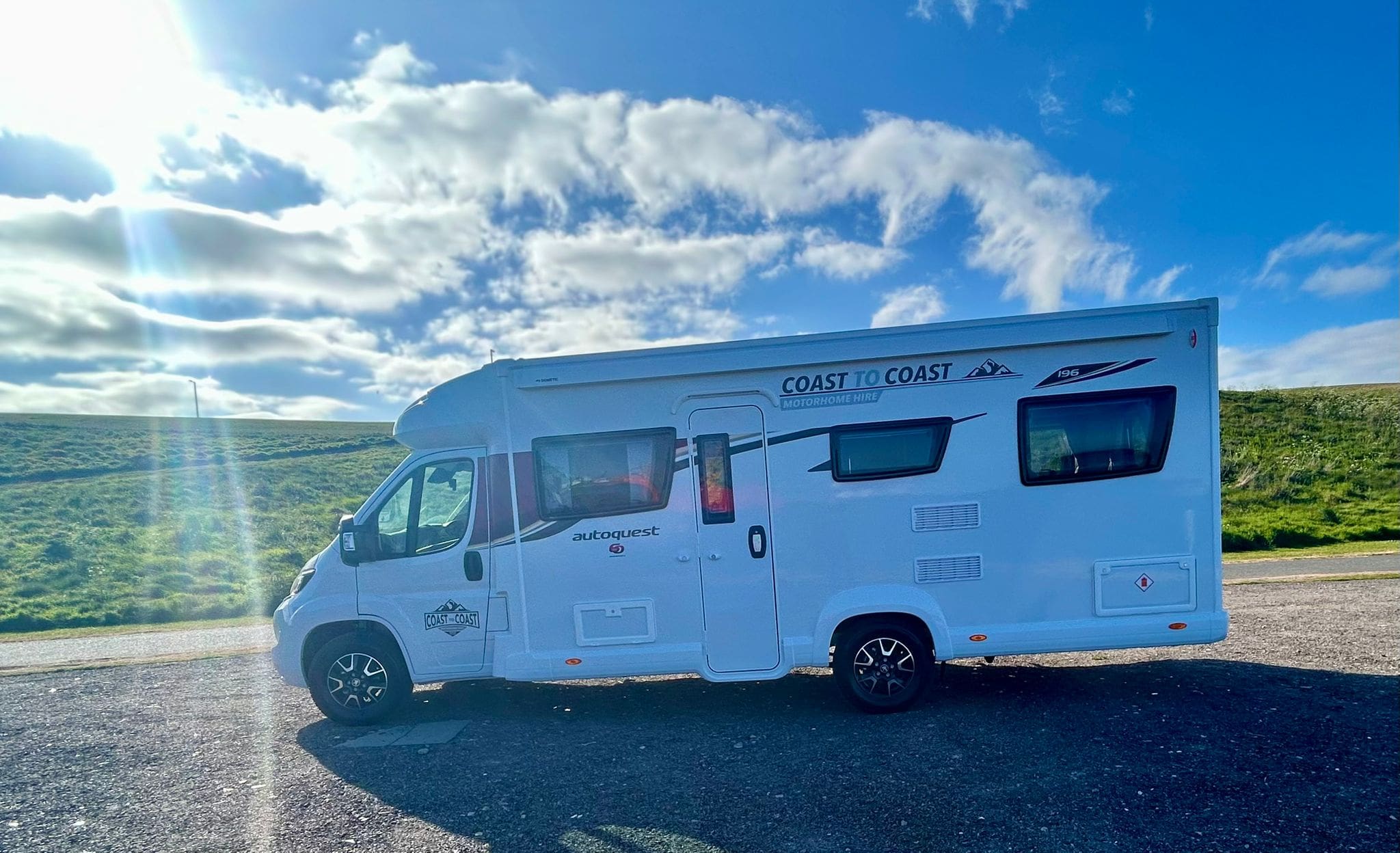 A white motorhome labeled Coast to Coast is parked on a paved area in Peterhead, Scotland, under a bright blue sky with sun rays and scattered clouds, with a grassy hill in the background.