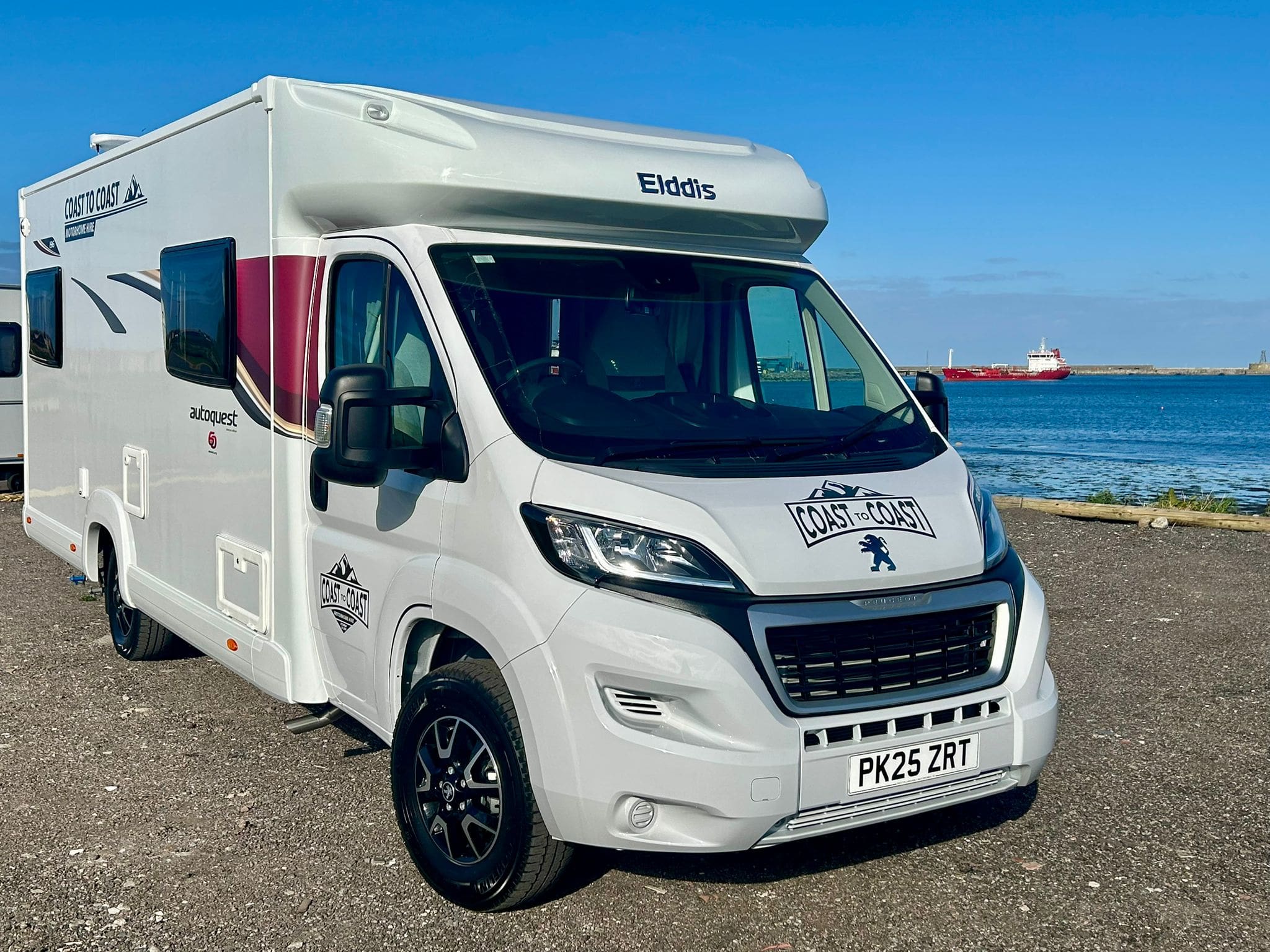 A white Elddis Coast to Coast motorhome is parked on a paved area by the sea in Peterhead, Scotland, with a clear blue sky and a red cargo ship in the background.
