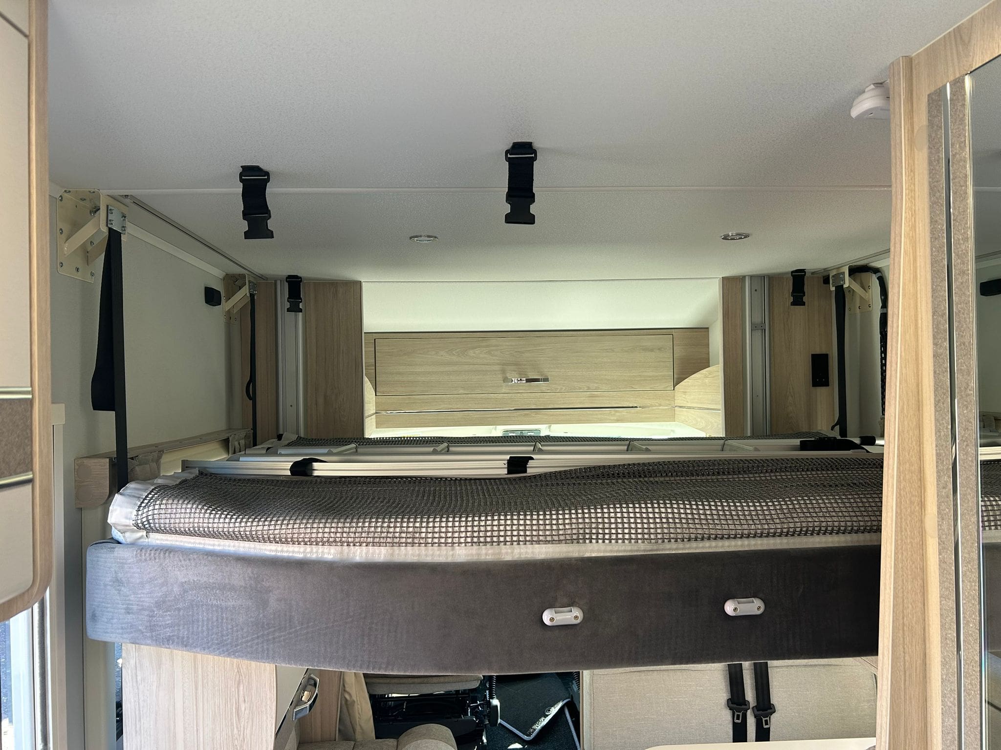 Interior of a rental motorhome in Scotland, featuring a raised bed platform with a rolled-up mattress, light wood cabinets, and storage above. Ceiling straps and modern fixtures are visible, with natural light entering from the left side.