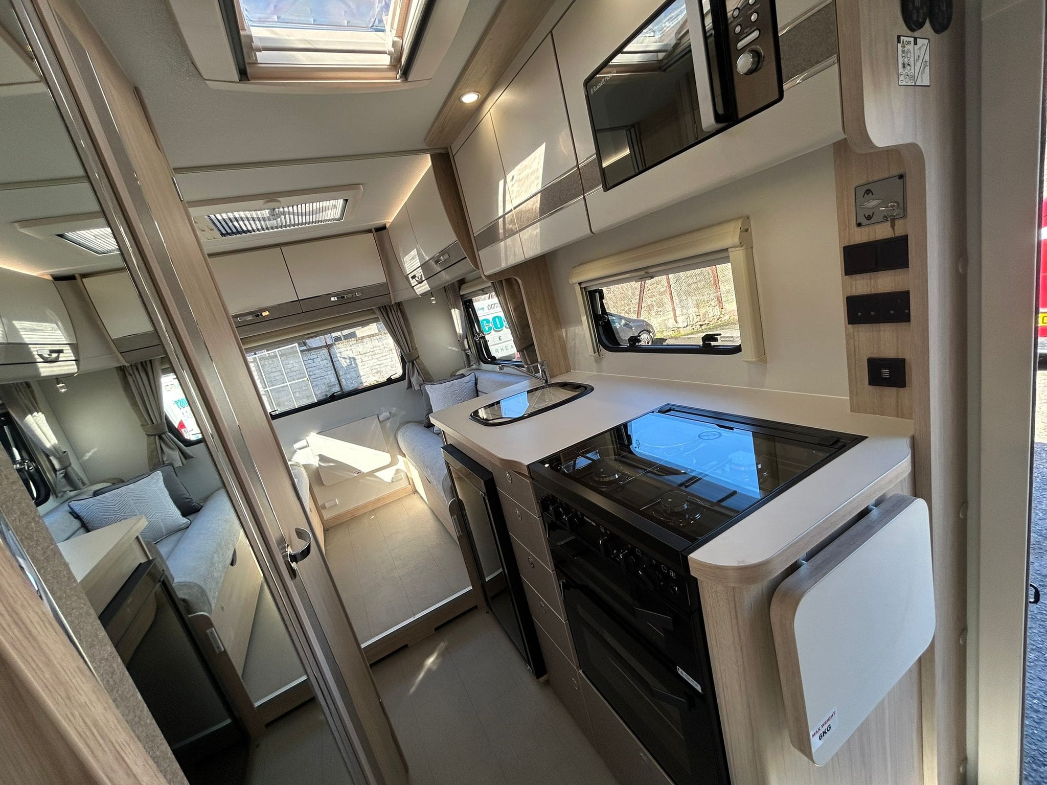 Modern campervan interior in Scotland, perfect for rental, featuring a compact kitchen with stove, oven, sink, microwave, and storage cabinets on the right. A seating area with windows and overhead storage basks in natural light.