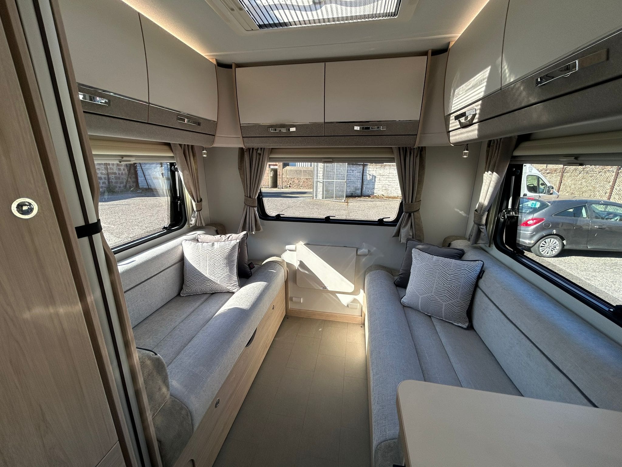 Interior of a modern caravan with two light gray sofas, matching cushions, large windows with curtains, overhead cabinets, and wood accents. Daylight streams in, and a car is visible outside through the window.