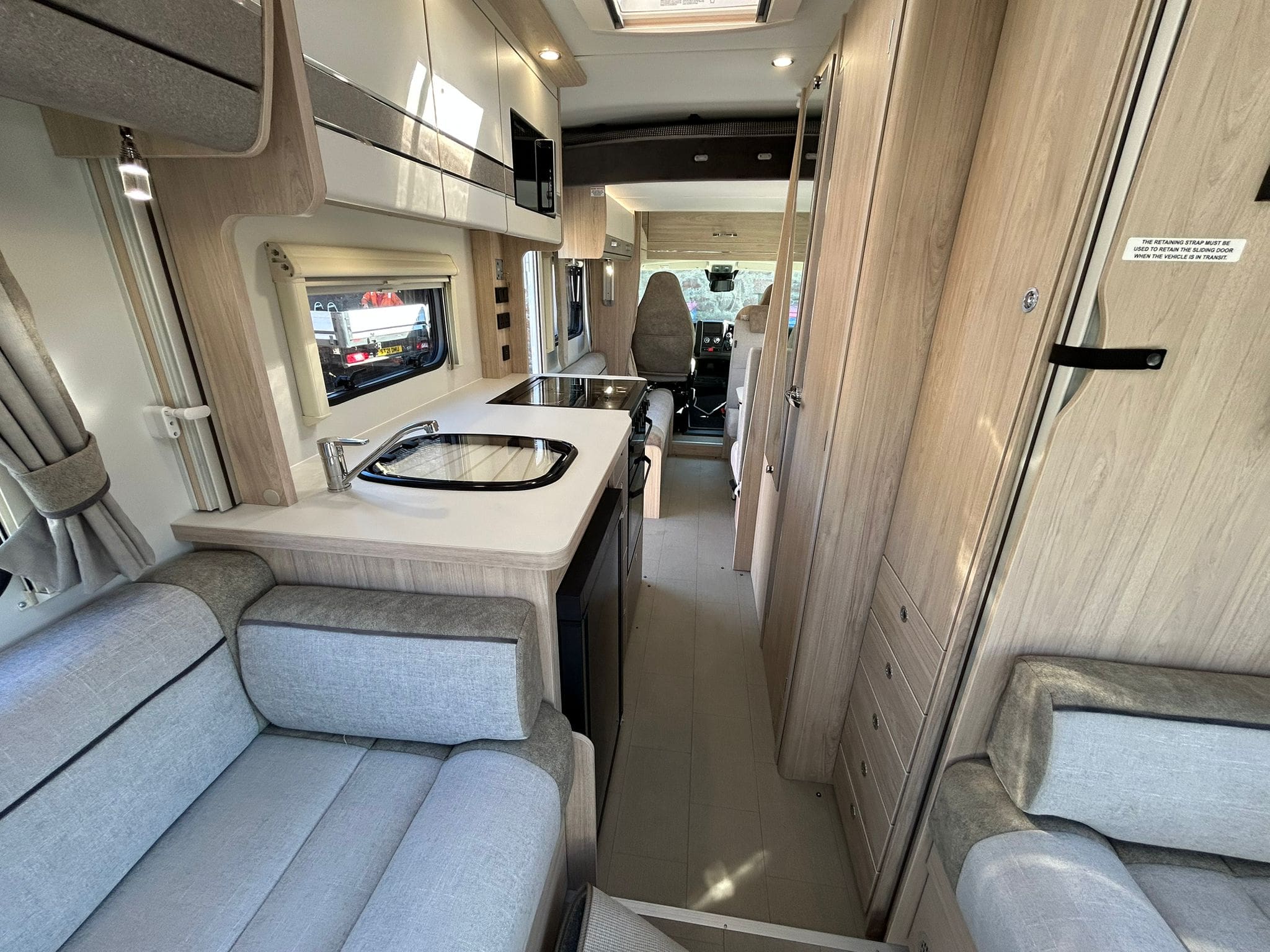 Interior of a modern motorhome rental featuring light wood cabinets, a compact kitchen with stove and sink, gray cushioned seating, and the driver’s area at the front—ideal for exploring Scotland. Natural light brightens the space.