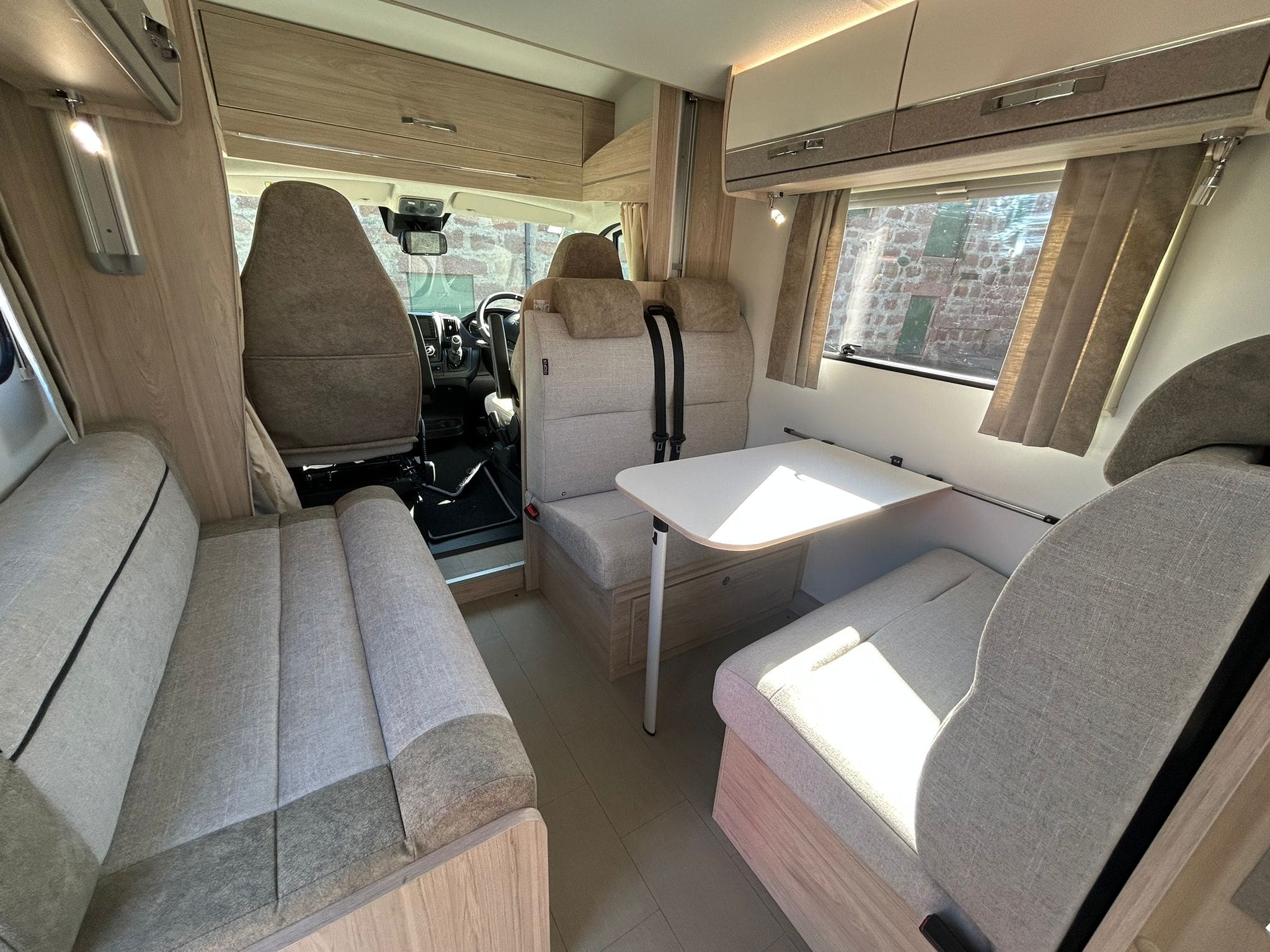 Interior of a modern motorhome near Peterhead, Scotland, featuring beige and gray seating, a small dining table, windows with curtains, and storage cabinets above. Driver and passenger seats face the living area.