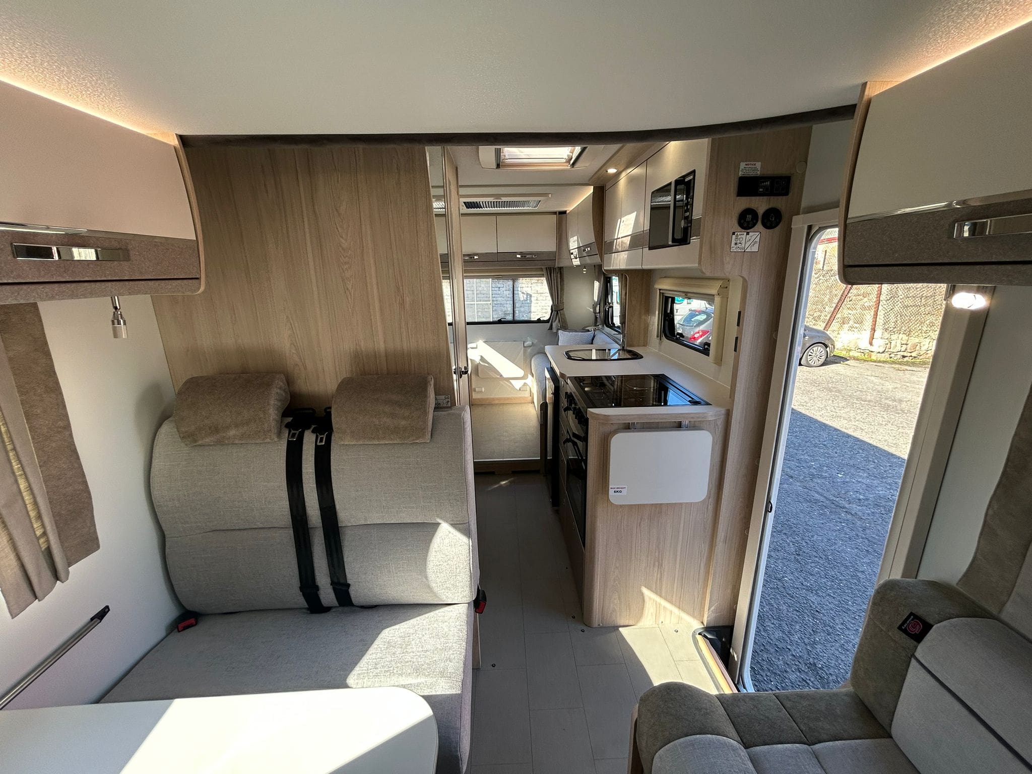 Interior of a modern motorhome rental with light wood and beige decor, featuring seatbelted seats, a dining table, kitchen area with sink and stove, plus a view toward the driver’s cab and open door—perfect for exploring Scotland.